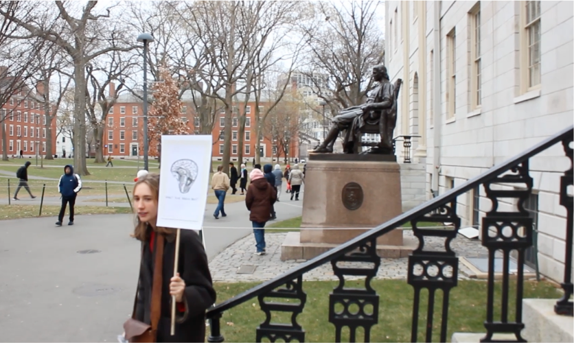 Student marching through Harvard Yard with a &lsquo;want your brain back?&rsquo; sign